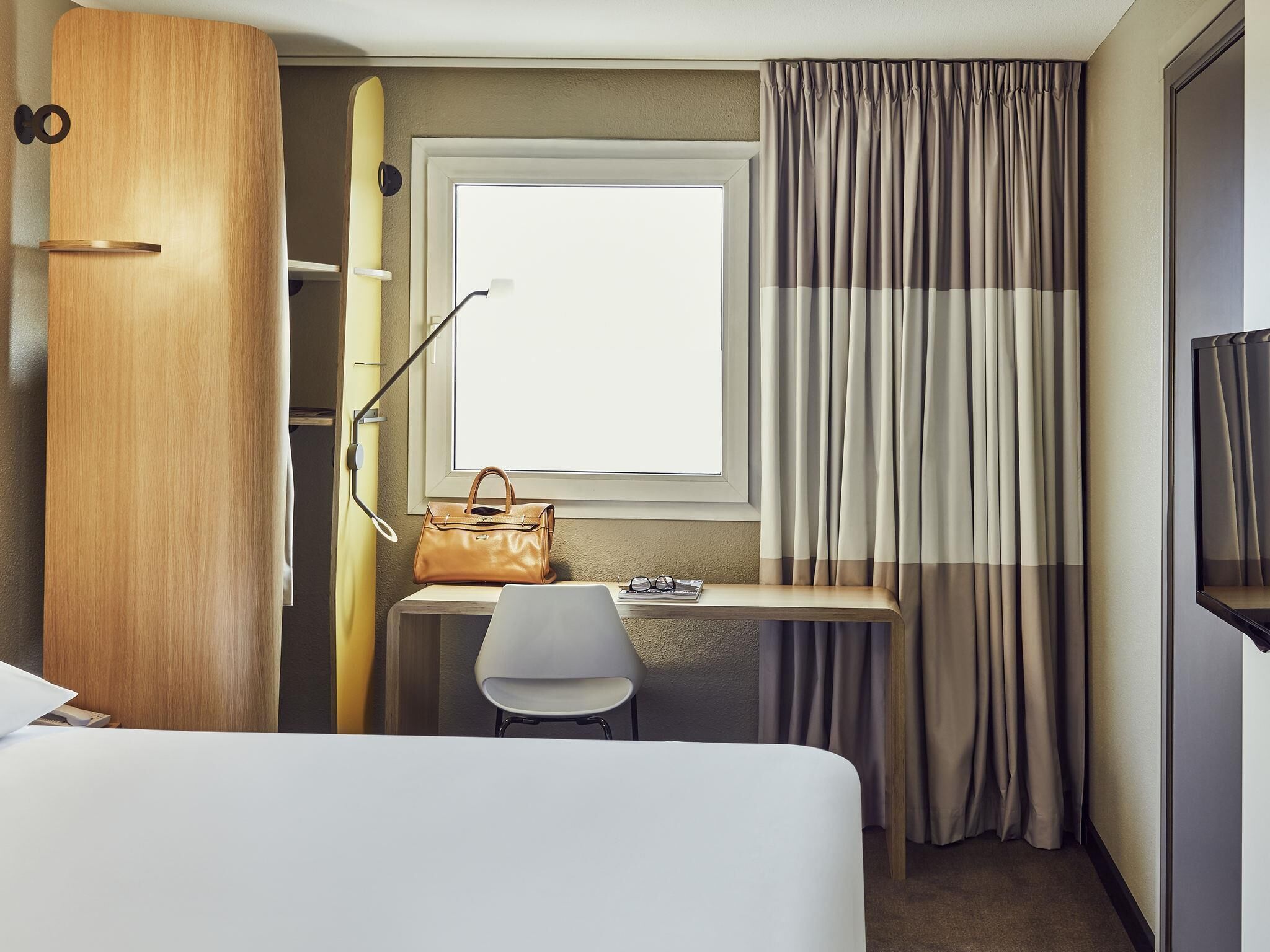 Een hotelkamer met een bed, een bureau met stoel, een raam met gordijnen, een surfplank en een handtas.