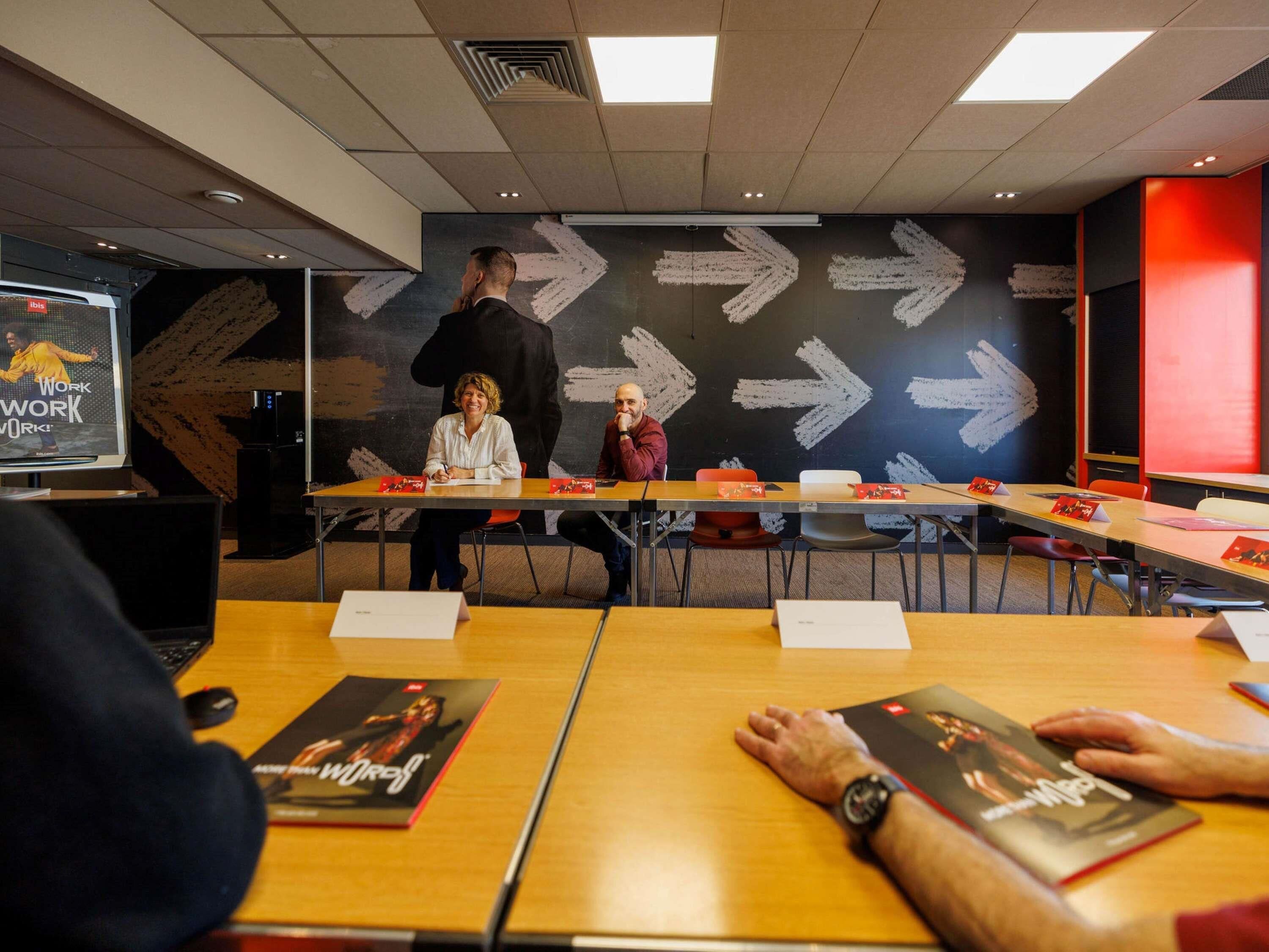 Una sala de conferències amb taules, cadires i una pantalla gran que mostra una imatge.