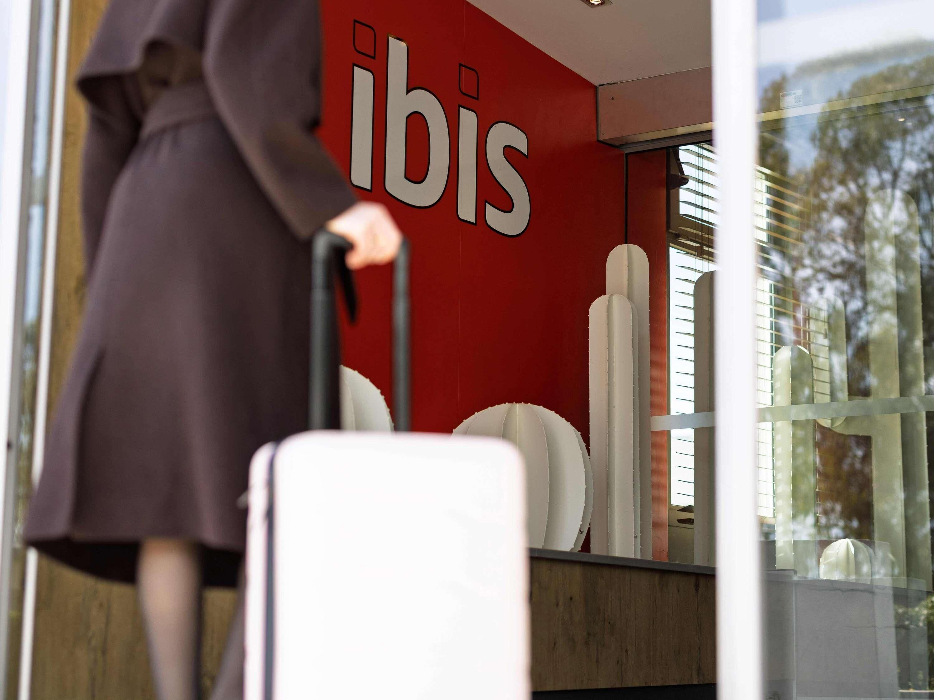 A person with a suitcase entering a building with a red wall and the word 'ibis' on it.