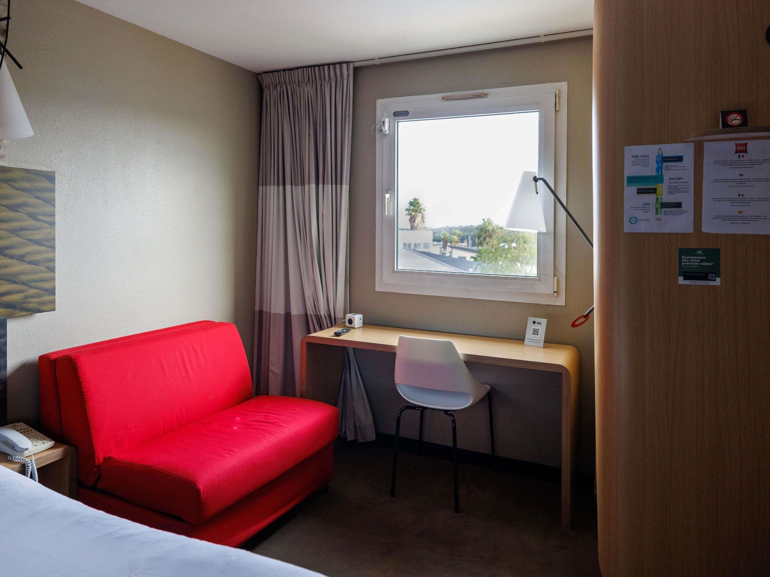 A hotel room with a red sofa, a desk with a chair, a window with curtains, and a lamp.