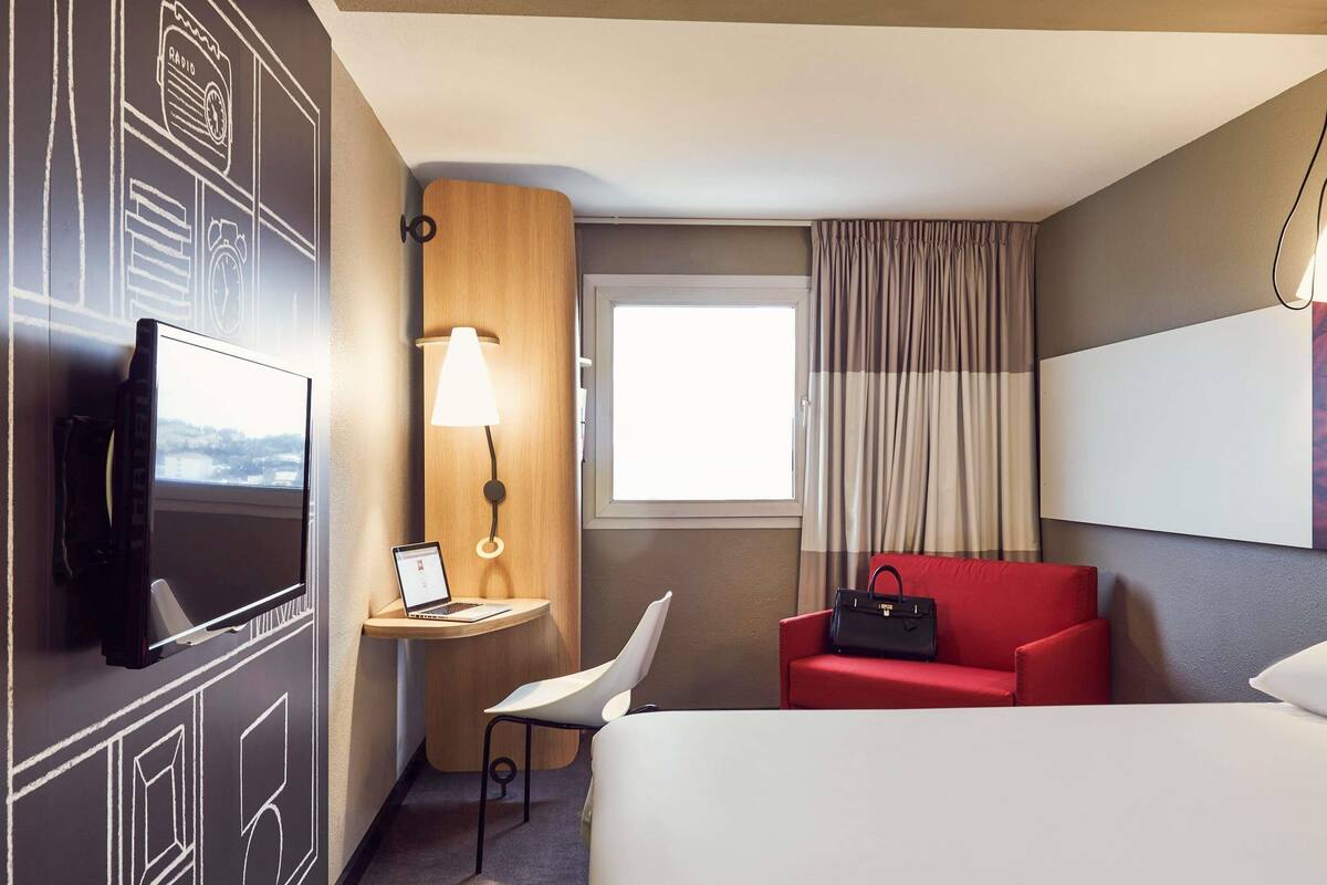 A hotel room with a bed, a desk with a laptop, a TV, a red chair, a lamp, and a window with curtains.