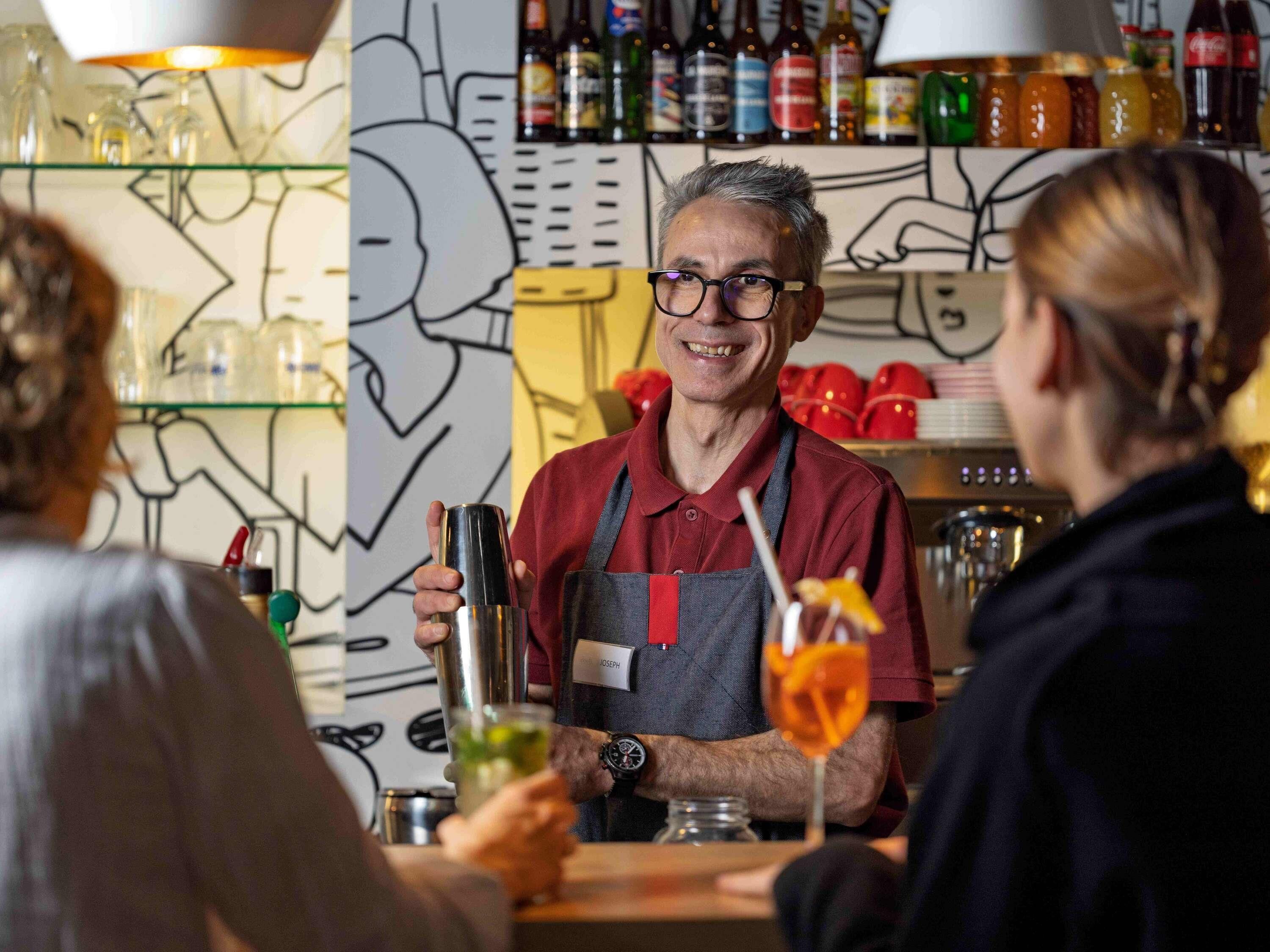 A bar setting with a bartender, shelves with bottles, and a wall with a line drawing.