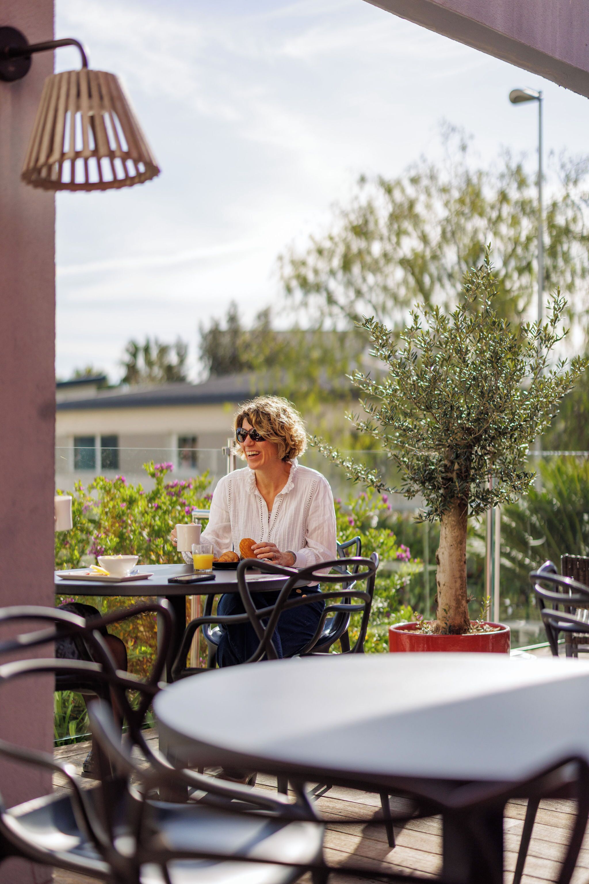 Una persona sentada en una mesa al aire libre, con una bebida y comida, rodeada de sillas y una planta en maceta.