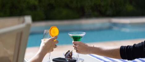Two people toasting with drinks at a poolside table with a smartphone and sunglasses.
