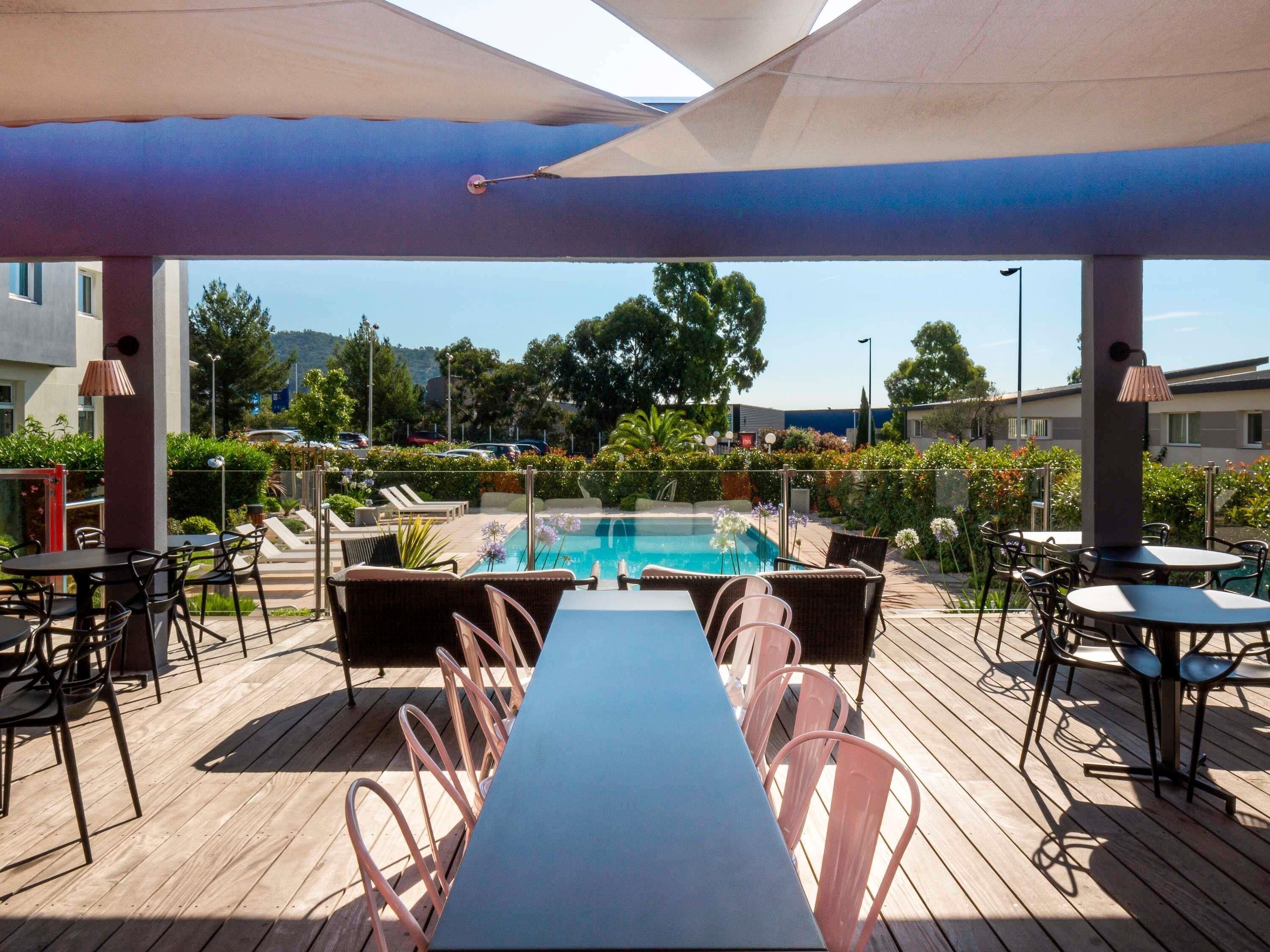 An outdoor pool area with a long table and chairs, surrounded by umbrellas and potted plants.