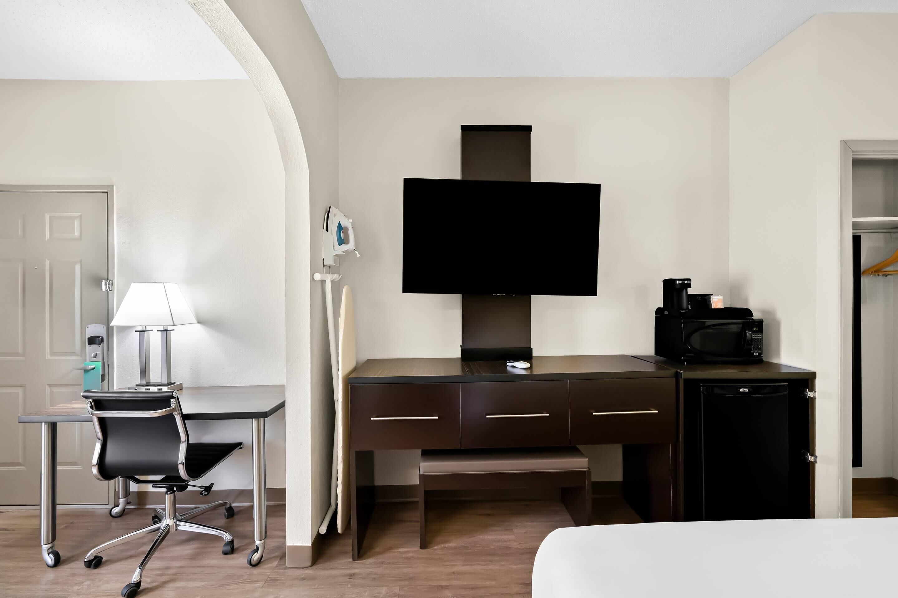 A hotel room with a desk, chair, television, microwave, and a small refrigerator.
