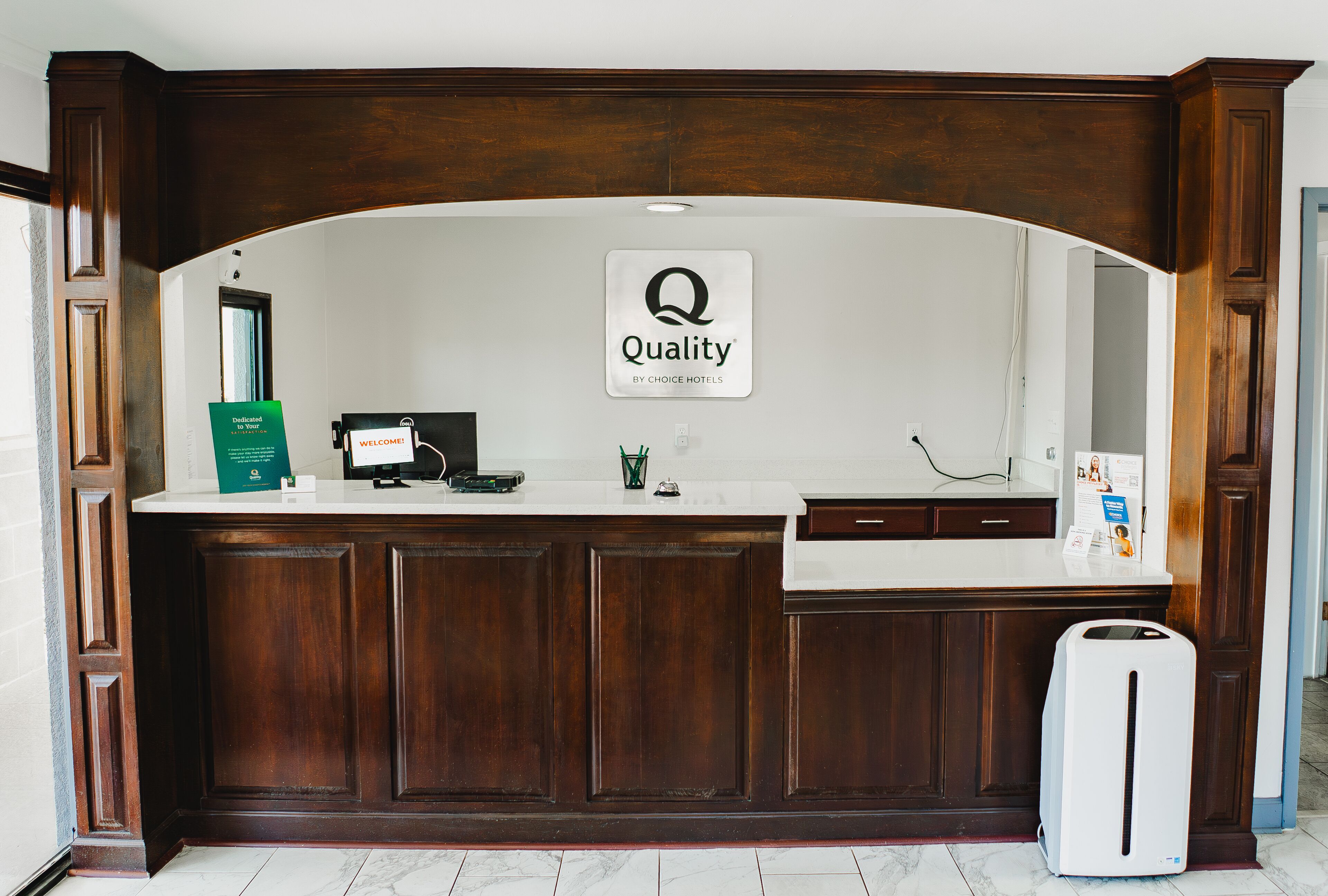 Un mostrador de recepción con un cartel que dice "Quality by Choice Hotels", una computadora, un letrero verde y un cubo de basura blanco.