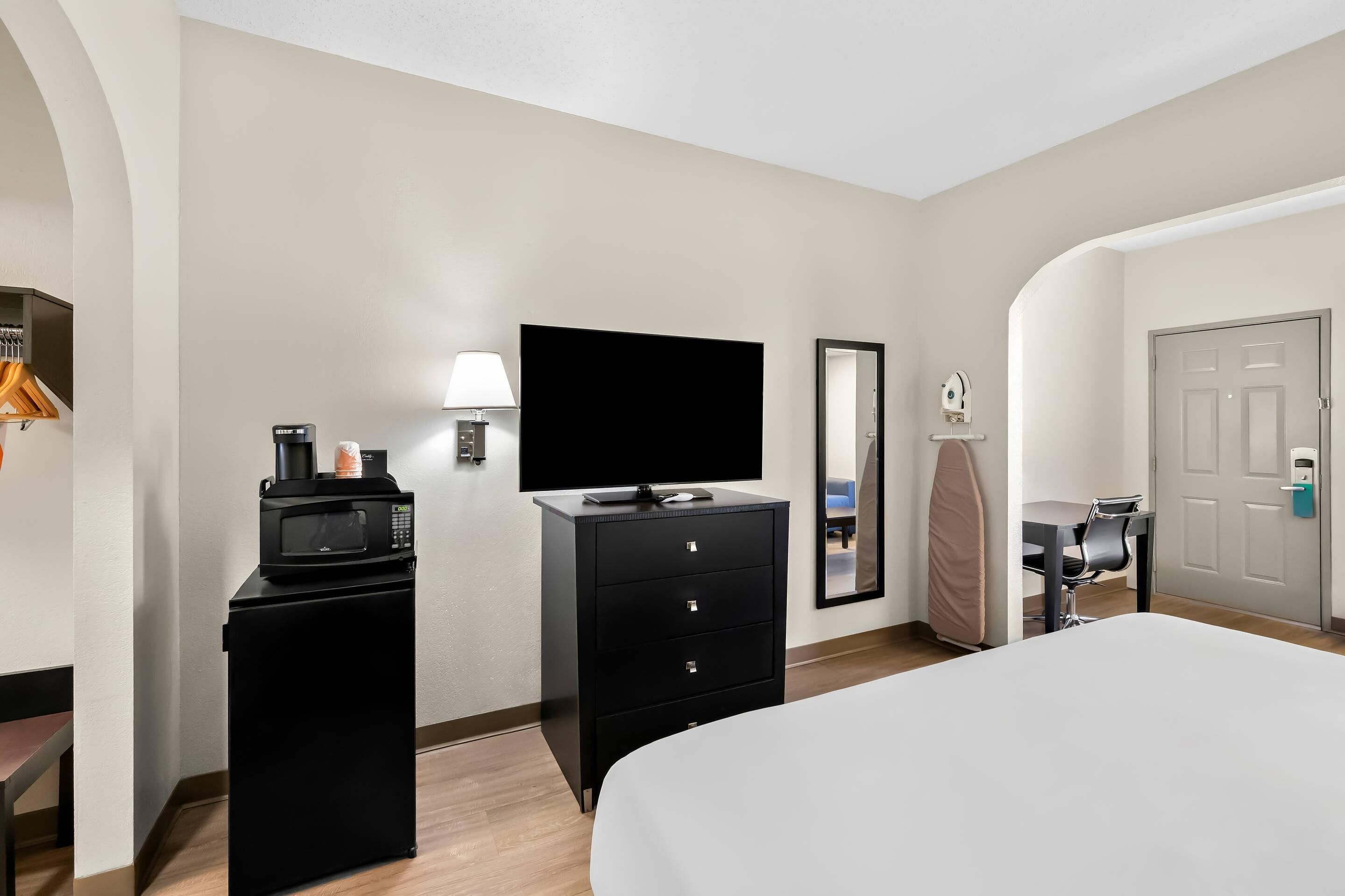 A hotel room with a bed, a desk with a chair, a black minibar, a flat-screen TV, a mirror, and a door.