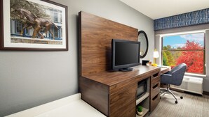 Una habitación de hotel moderna con un escritorio de madera, un televisor de pantalla plana, un sillón cómodo y un ventanal con vistas a árboles.