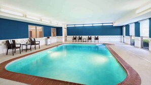Una piscina interior con borde de azulejos, rodeada de sillas y mesas, y ventanas amplias a un lado.