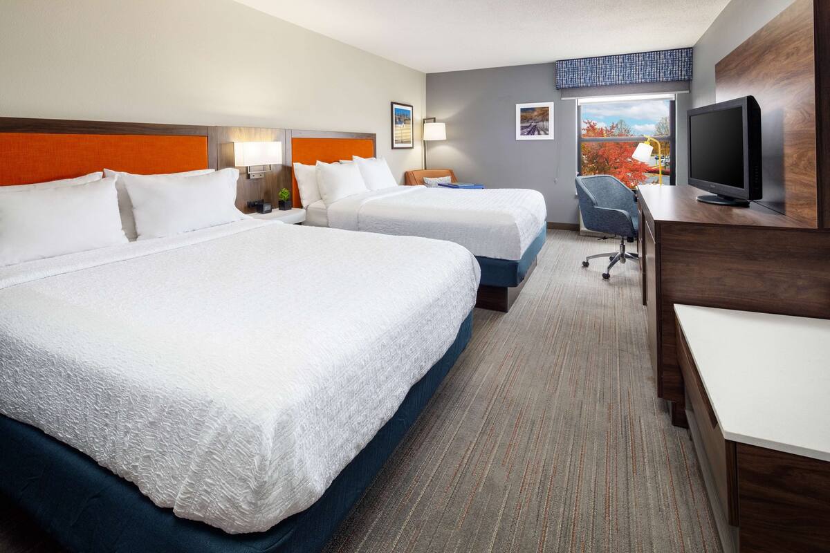 A hotel room with two beds, a desk, a television, and a window with a view of autumn foliage.