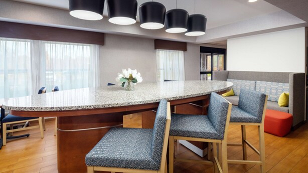 Una cocina moderna con una isla central, taburetes de bar, iluminación colgante y un sofá gris con un reposapiés rojo.