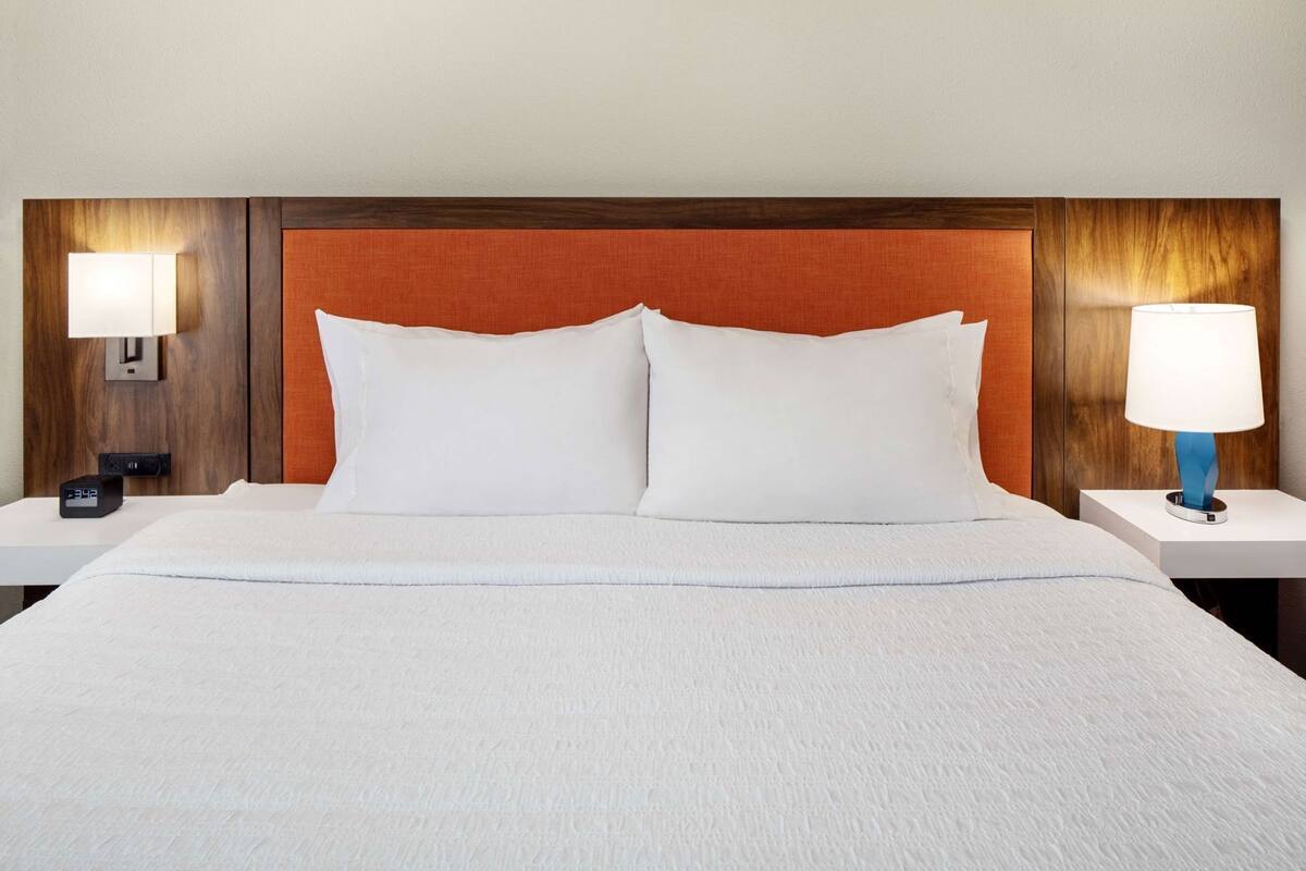 A hotel room with a large bed, two bedside tables, a digital clock, and a wall-mounted lamp.