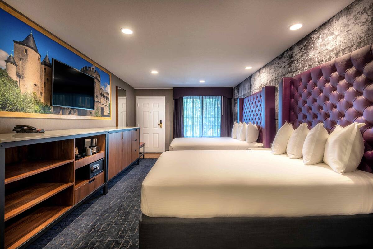A hotel room with a large flat-screen TV mounted on the wall, a bed with a tufted headboard, a wooden desk with a chair, and a window with curtains.