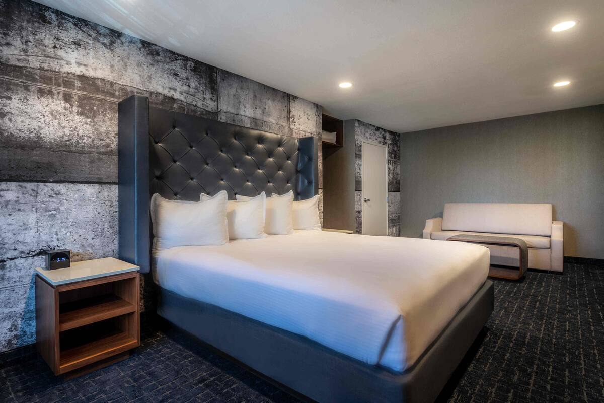 A modern hotel room with a large bed, a wooden nightstand, a seating area, and a textured wall.