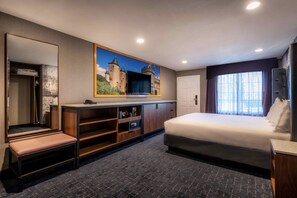 A modern hotel room with a large bed, a built-in bench, a wardrobe, a mirror, and a painting of a castle on the wall. - Anaheim Camelot Inn & Suites (Anaheim)