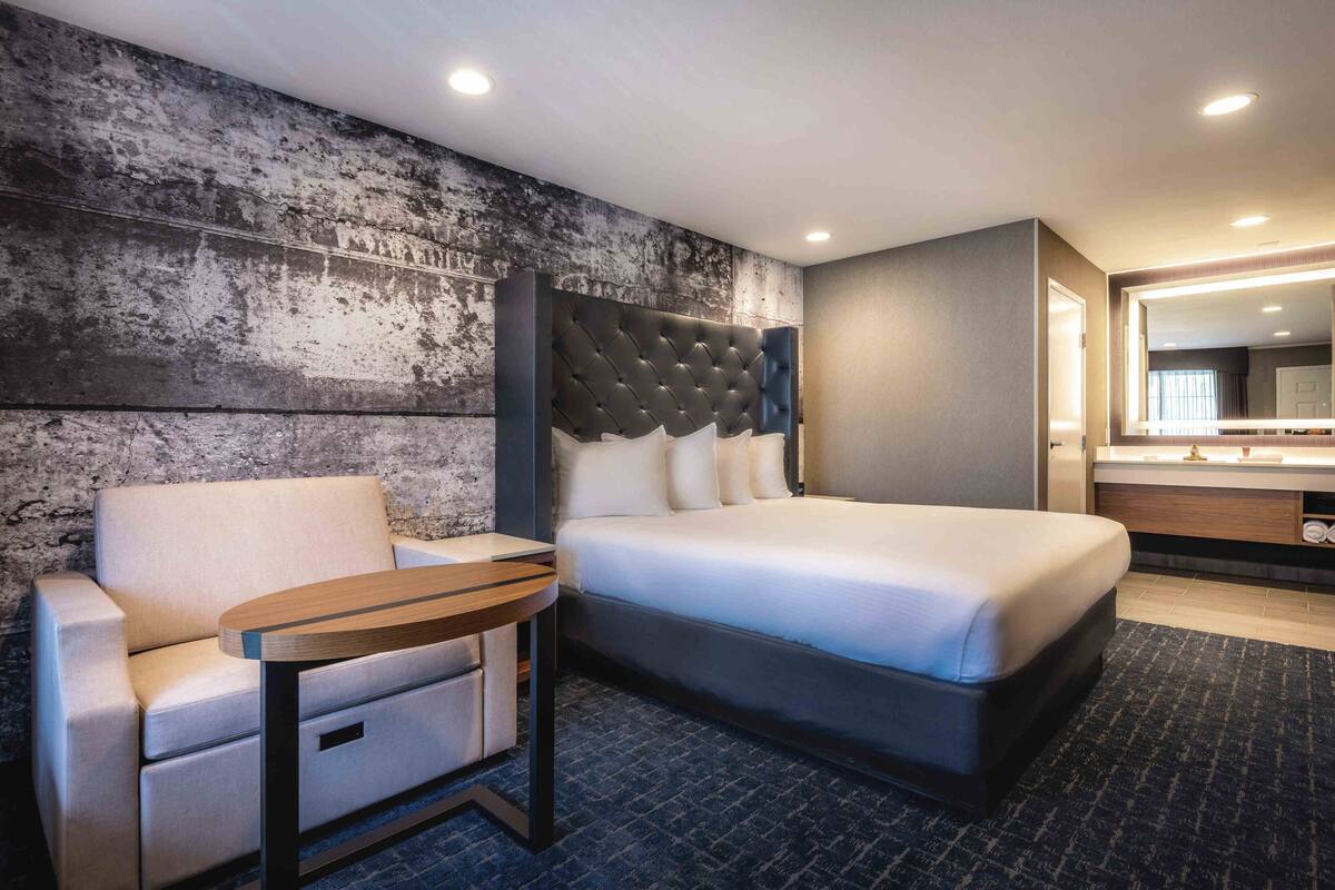 A hotel room with a bed, a chair, a small table, a mirror, and a bathroom with a sink and mirror.