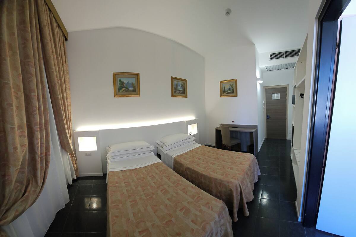 Une chambre d’hôtel avec deux lits, un bureau et une chaise. Il y a deux tableaux au mur, une fenêtre avec des rideaux et une porte qui donne sur une autre pièce.
