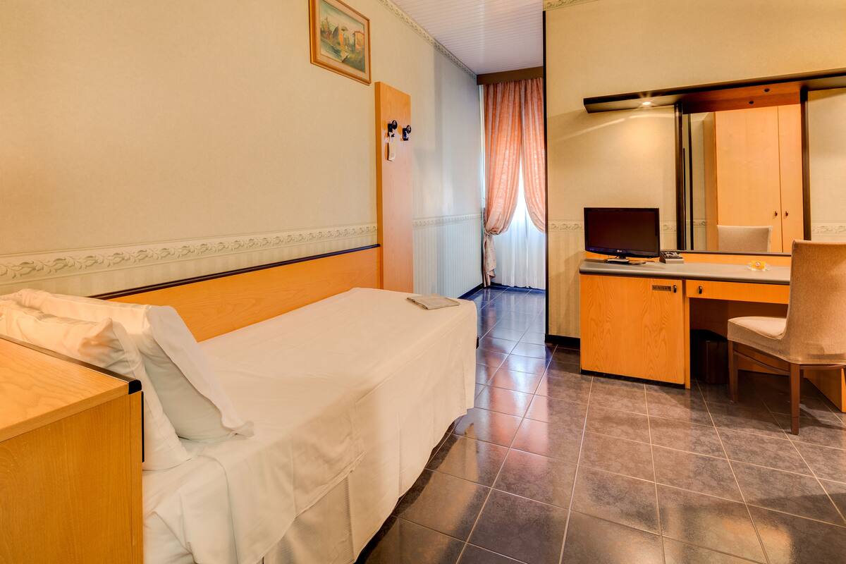 Une chambre d’hôtel avec un lit simple, un bureau avec un ordinateur, une chaise et un grand miroir.