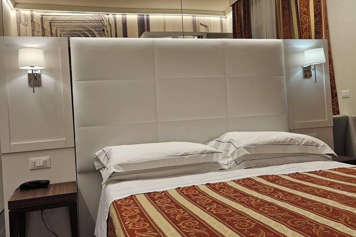 Une chambre d’hôtel avec un grand lit, une table de chevet avec une lampe, une prise électrique et un petit bureau avec une télécommande.