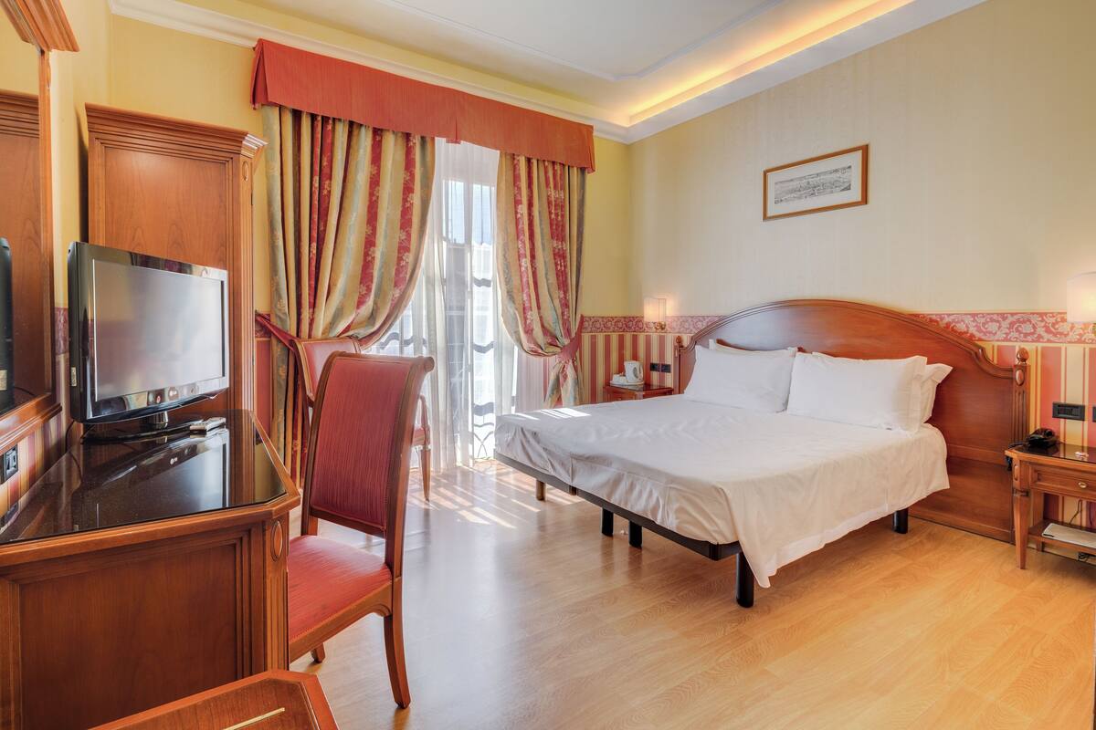 Une chambre d’hôtel avec un lit, un bureau avec une télévision, une chaise et une fenêtre avec des rideaux.