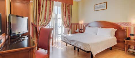 A hotel room with a bed, a desk with a television, a chair, and a window with curtains.