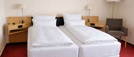 A double bed with white linens, two bedside tables with lamps, a chair, and a red carpet.
