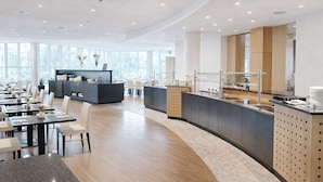A modern hotel dining area with a central buffet station, multiple seating arrangements, and large windows providing natural light. - Hotel Dresden Neustadt (Dresden)