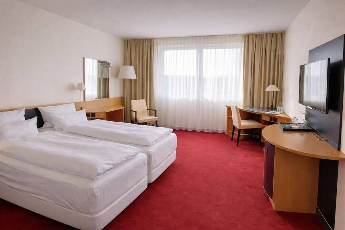 Ett hotellrum med två sängar, ett skrivbord, en stol, en TV och ett stort fönster med gardiner.