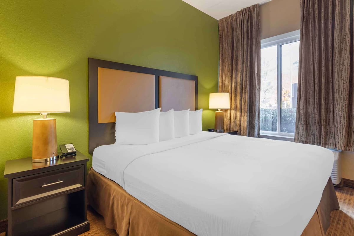 Ein Hotelzimmer mit einem großen Bett, zwei Nachttischen mit Lampen, einem Beistelltischchen mit Telefon und einem Fenster mit Vorhängen.