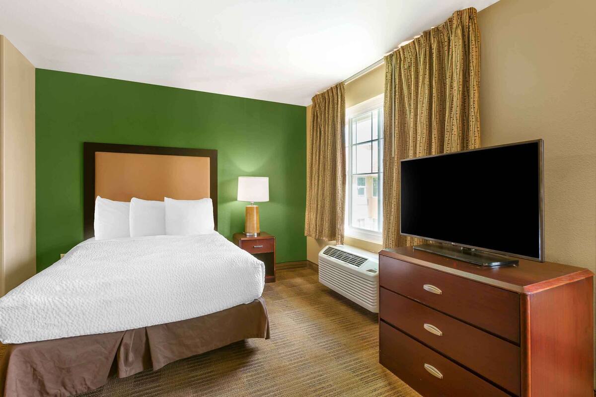 Ein Hotelzimmer mit einem großen Bett, einem Flachbildfernseher, einem Nachttisch, einer Lampe und einem Fenster mit Vorhängen.