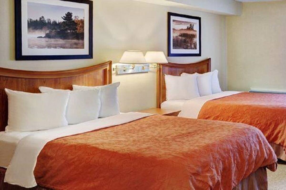 A hotel room with two beds, each with white linens and a red bedspread. There are two framed pictures on the wall, a nightstand with a lamp, and a window with curtains.