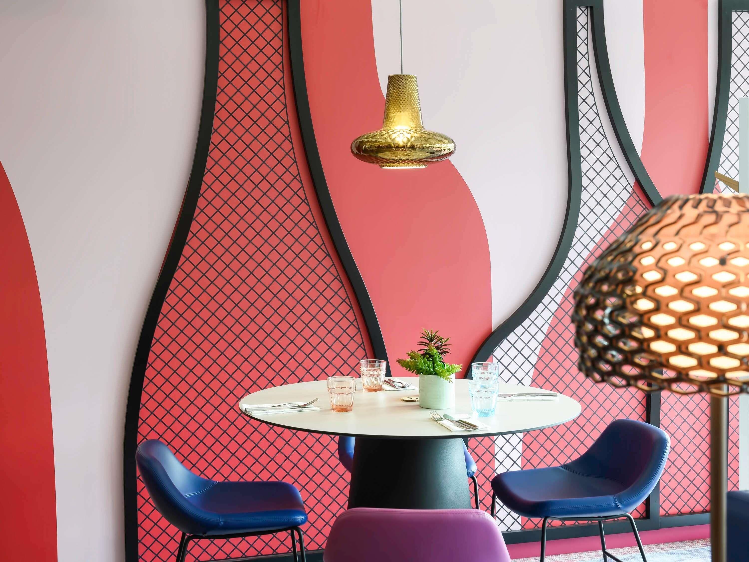 Een eetgedeelte met een ronde tafel, twee blauwe stoelen, een kleine potplant en een hangende gouden hanglamp.