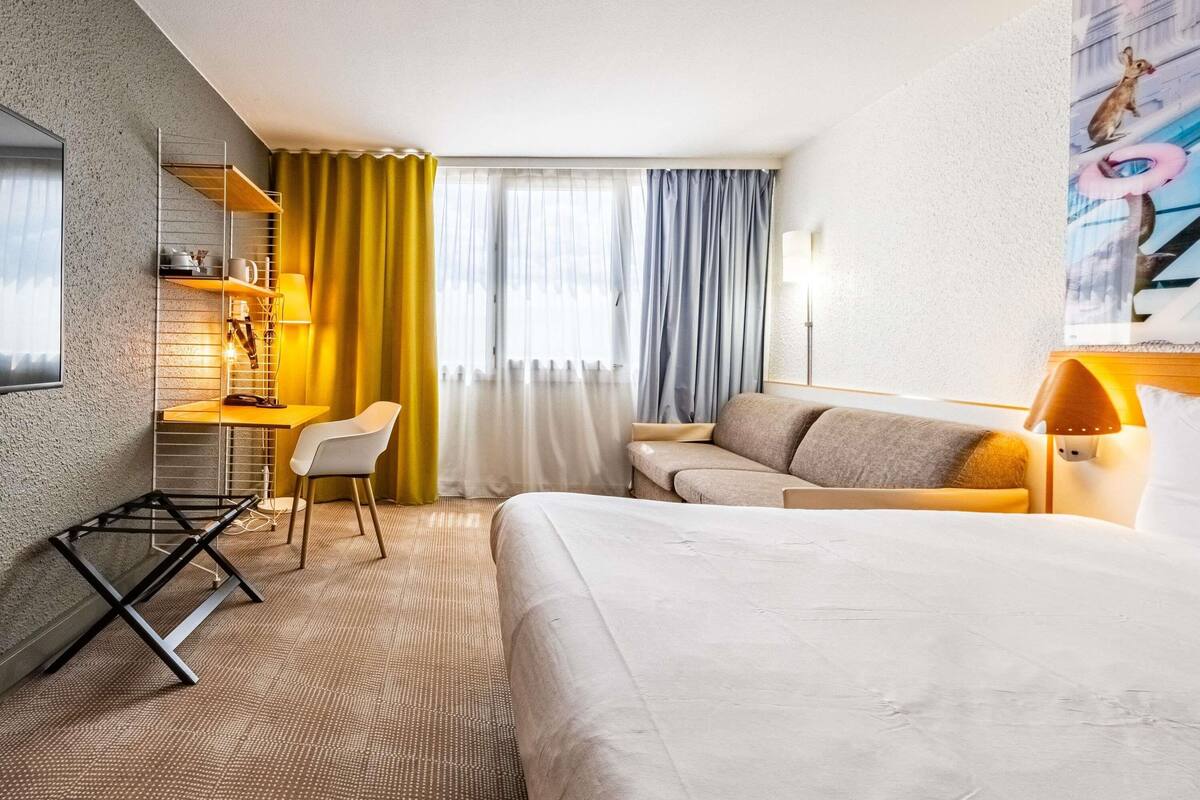 A hotel room with a bed, a desk, a chair, a sofa, and a window with curtains.
