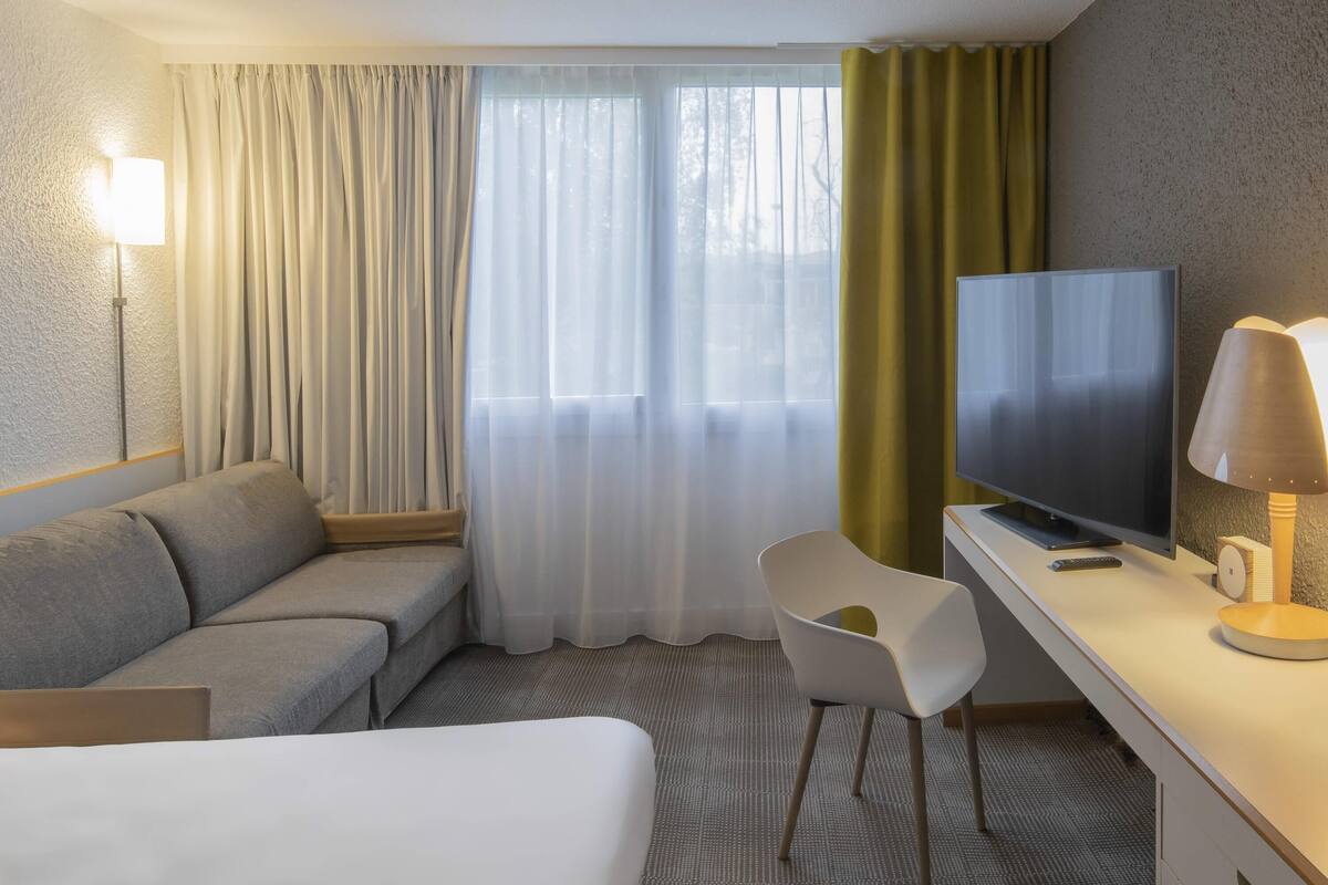 A hotel room with a bed, a sofa, a desk with a television, a chair, and a lamp.