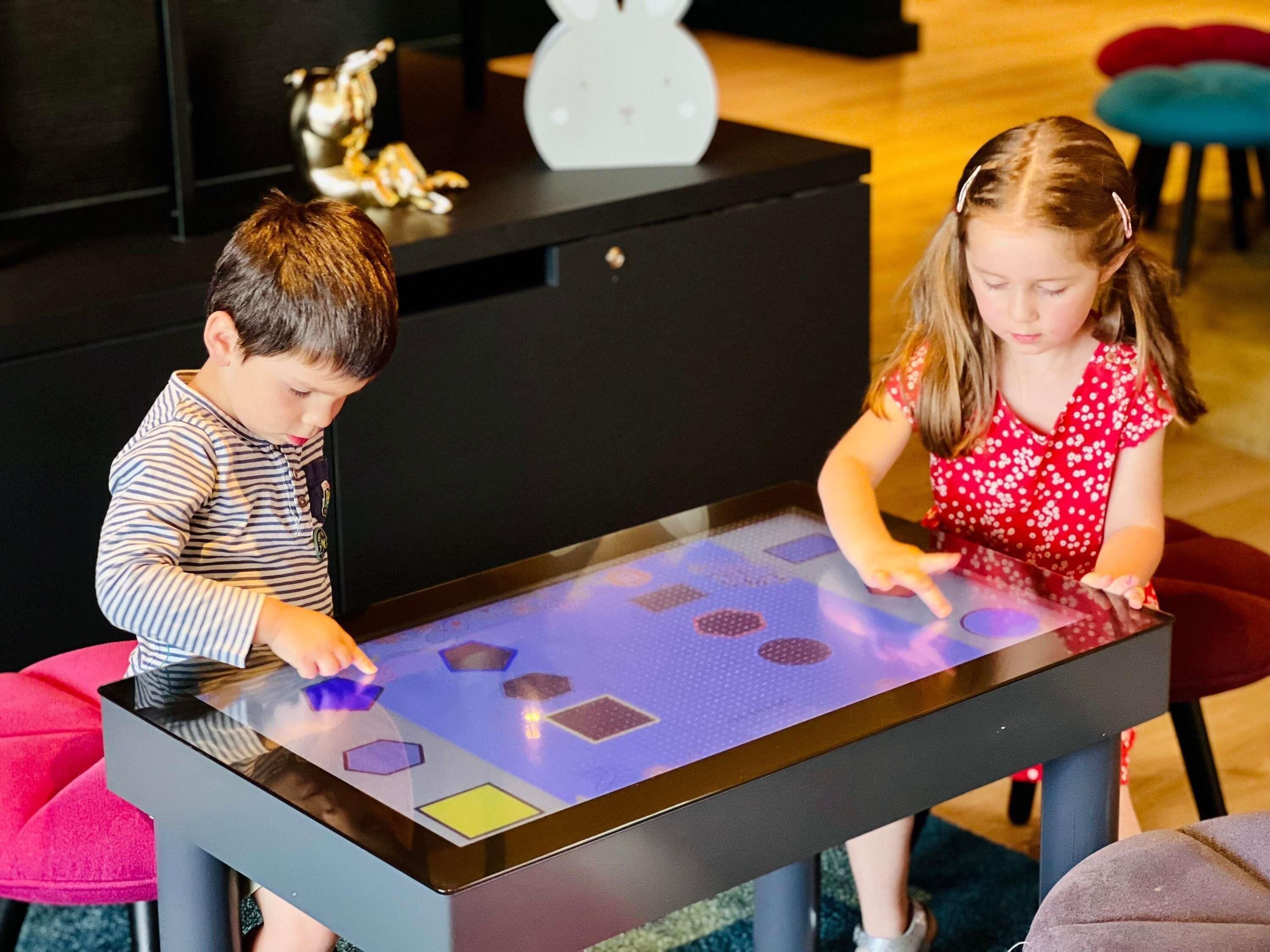 Deux enfants jouent sur une table interactive numérique dans une pièce où se trouvent un meuble noir et des chaises colorées.