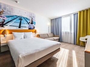 A hotel room with a bed, a sofa, a chair, a painting on the wall, and a window with curtains. - Novotel Dijon Sud (Marsannay-la-Cote)
