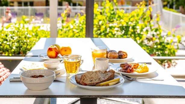 A breakfast table with various food items and a glass of juice, set outdoors with a view of greenery and buildings in the background.