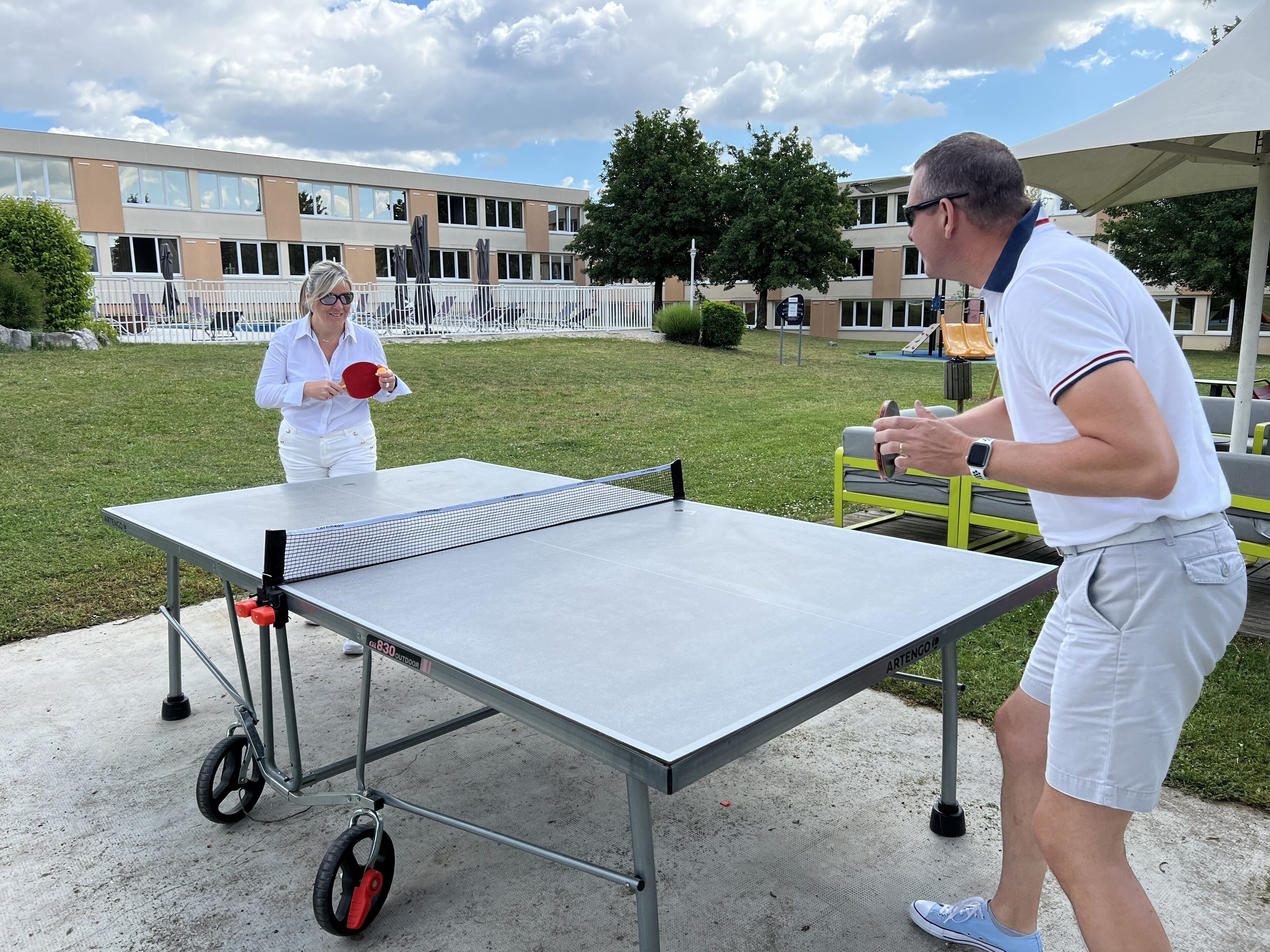 Mesa de ping pong al aire libre instalada en un área verde con un edificio al fondo.