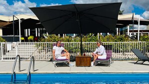Two people seated by a poolside with umbrellas, a pool, and a white fence in the background.