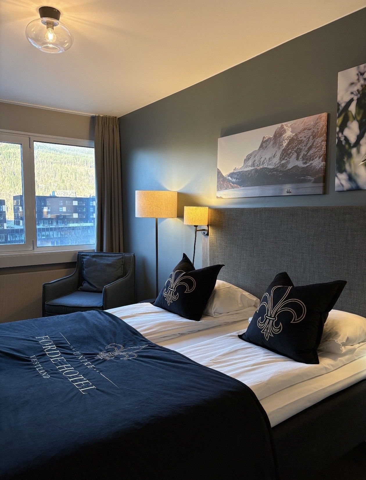Habitación de hotel con una cama, dos lámparas de noche, una silla, una ventana con vistas a edificios y un cuadro en la pared.