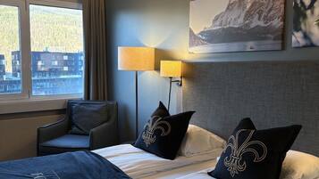 A hotel room with a bed, two bedside lamps, a chair, a window with a view of buildings, and a painting on the wall.