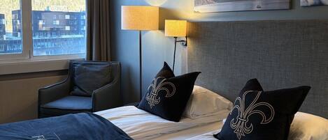 Habitación de hotel con una cama, dos lámparas de noche, una silla, una ventana con vistas a edificios y un cuadro en la pared.