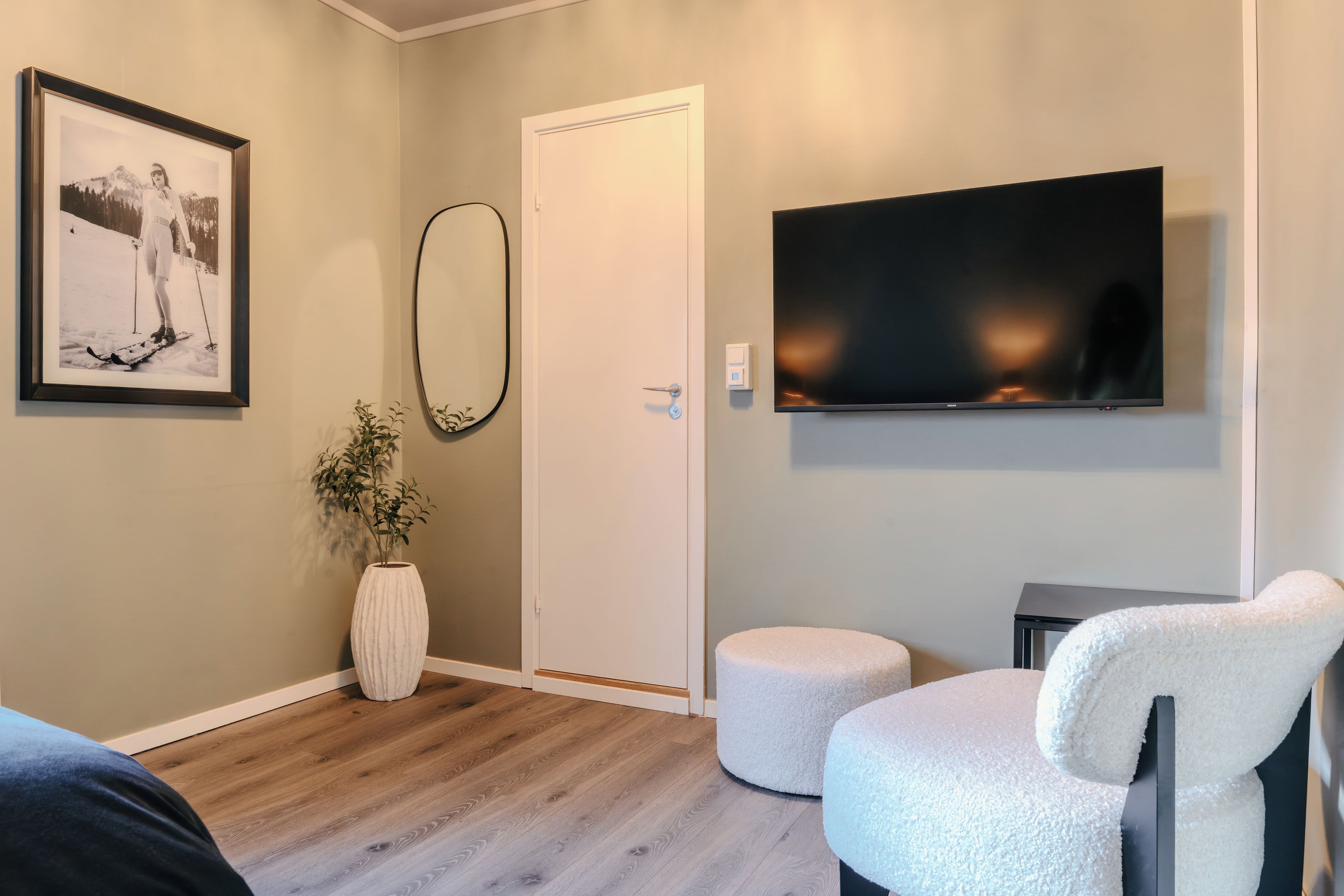 Habitació amb sòl de fusta, porta blanca, fotografia emmarcada, mirall gran, televisió de pantalla plana i dues butaques blanques.