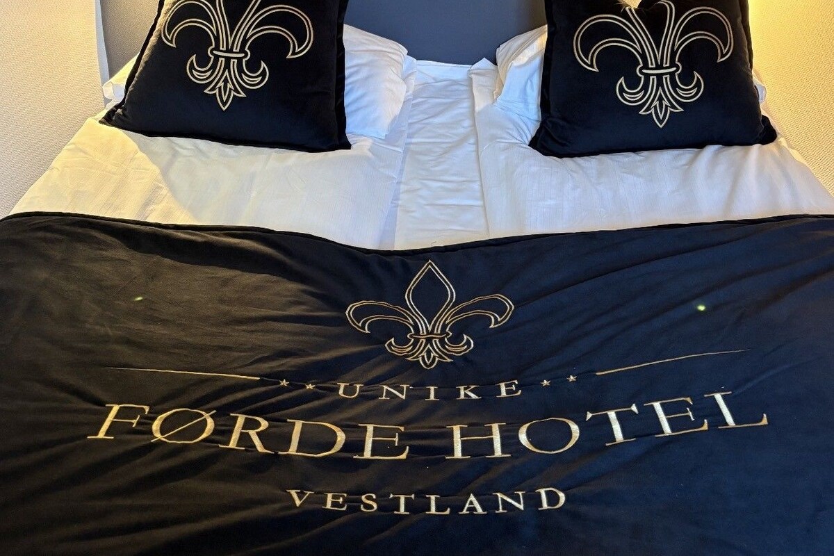 A hotel room with a bed, two pillows with fleur-de-lis designs, a dark bedspread with 'UNIKE FORDE HOTEL VESTLAND' text, and a wooden headboard.
