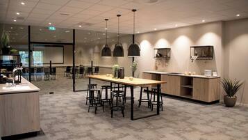 A modern office space with a long wooden table surrounded by chairs, a kitchenette, and a conference room visible through glass walls.