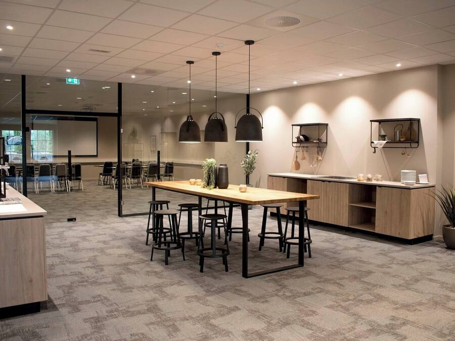 A modern office space with a long wooden table surrounded by chairs, a kitchenette, and a conference room visible through glass walls.