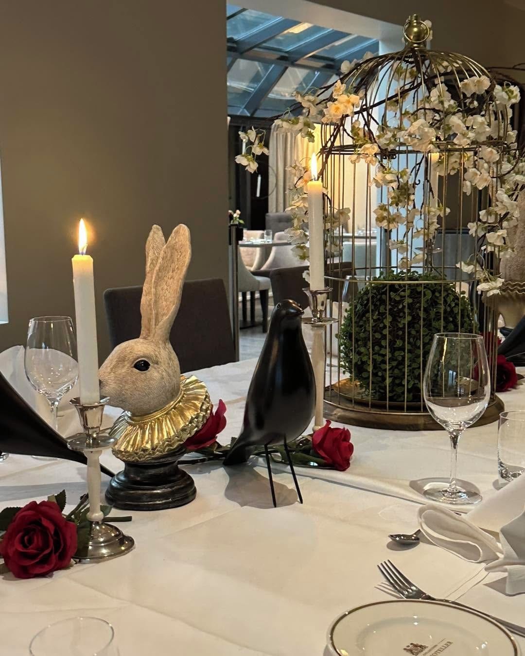 Una mesa de comedor dispuesta con una vela, una figurita de pájaro, una figurita de conejo y una jaula de pájaros con flores.