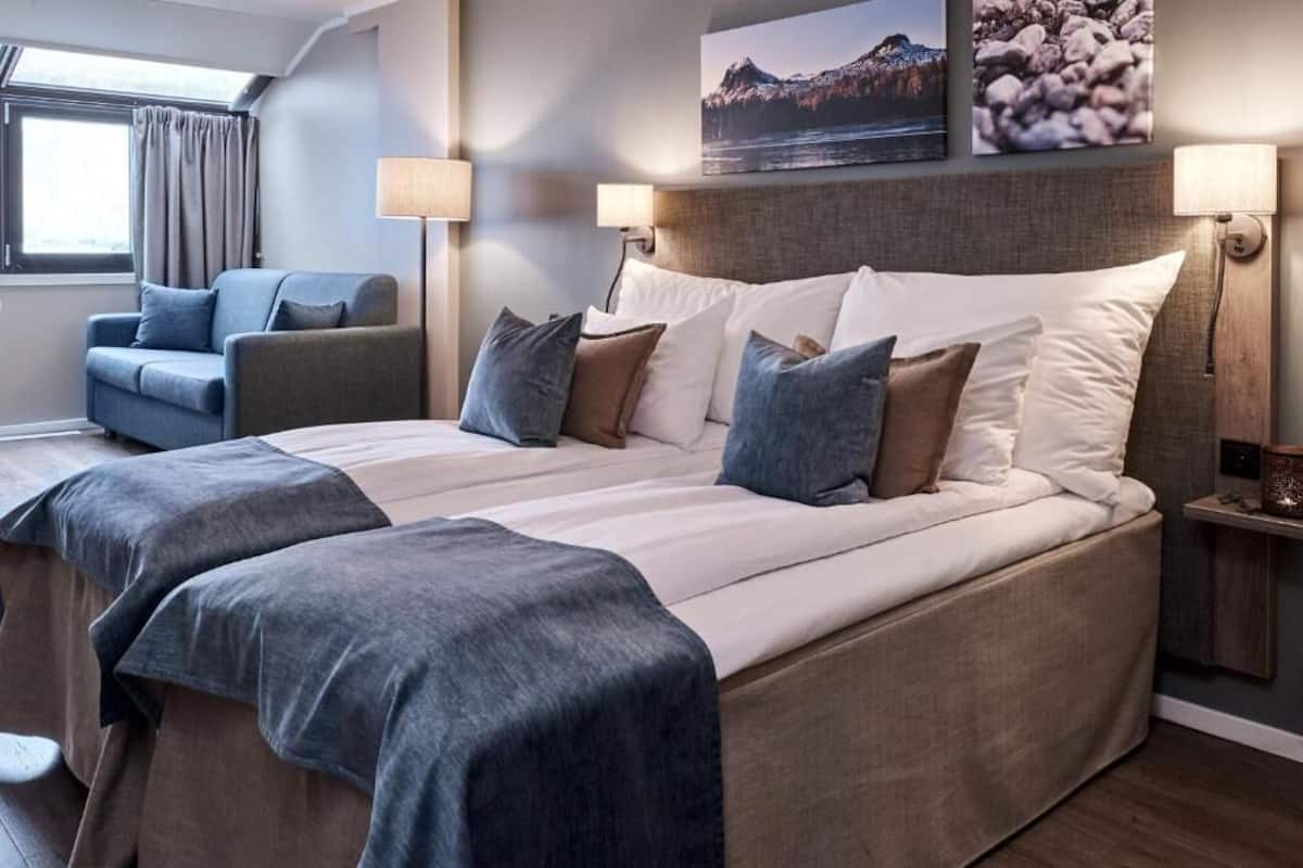 A double bed with a headboard, two bedside tables with lamps, a blue armchair, and a window with curtains.