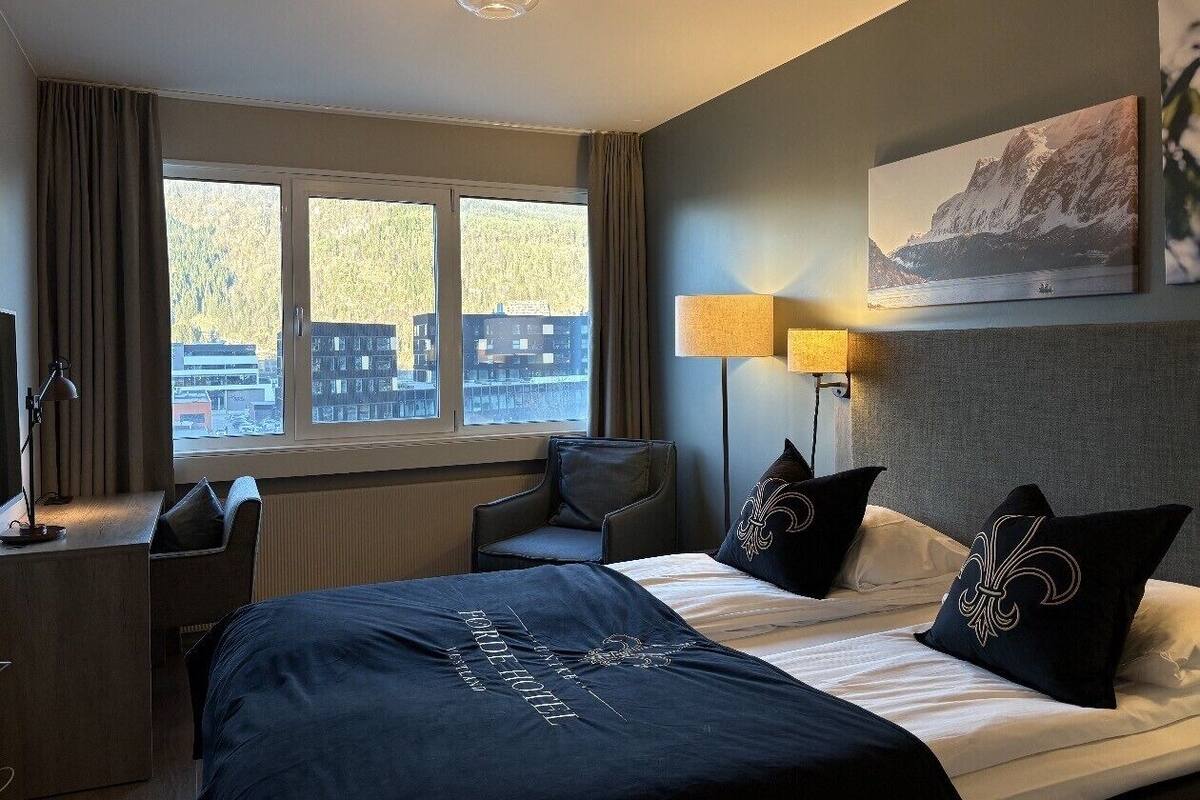 Habitación de hotel con cama, escritorio, silla, lámpara y vistas a edificios a través del ventanal.